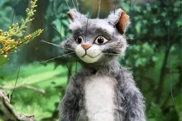 Eine graue Puppenkatze mit großen Augen und einem freundlichen Gesicht steht in einem grünen Wald. Die Katze hat eine weiche Fellstruktur und wirkt lebhaft.
