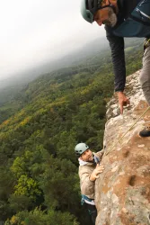 Zwei Kletterer am Felsen in einer grünen, bewaldeten Landschaft. Der eine Kletterer erreicht die Spitze, während der andere ihm Anweisungen gibt.