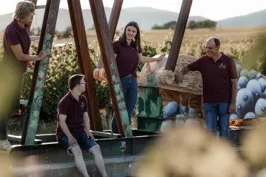 Eine Gruppe von vier Personen steht an einem Handwerkzeug, umgeben von Weinbergen. Sie tragen einheitliche lila Poloshirts und wirken freundlich und entspannt.