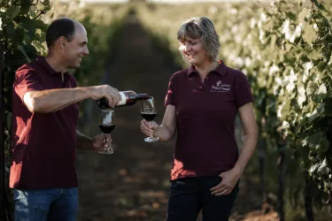 Ein Paar steht in einem Weinberg und genießt Wein. Der Mann schenkt der Frau ein Glas Rotwein ein.