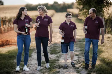 Eine Gruppe von vier Personen geht auf einem Weg durch Weinberge. Sie halten Flaschen und Gläser Wein in der Hand und tragen einheitliche T-Shirts.