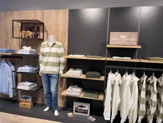 Ein moderner Bekleidungsstore mit einem Mannequin in einem gestreiften Pullover und Jeans. Im Hintergrund sieht man verschiedene gestapelte Kleidungsstücke und Shirts auf Regalen.