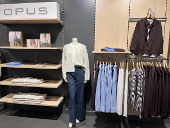 Ein Modegeschäft mit einer Schaufensterpuppe, die eine Damenoberbekleidung trägt. Regal mit Kleidung in verschiedenen Farben und Faltwaren im Hintergrund.