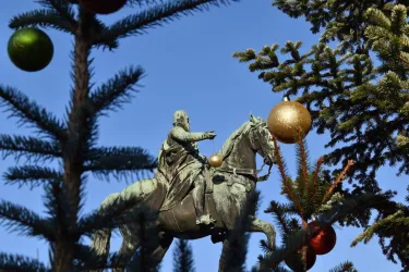 Eine Statue eines Reiters auf einem Pferd, umgeben von Tannenzweigen und Weihnachtskugeln. Der klare blaue Himmel bildet den Hintergrund der Szene.