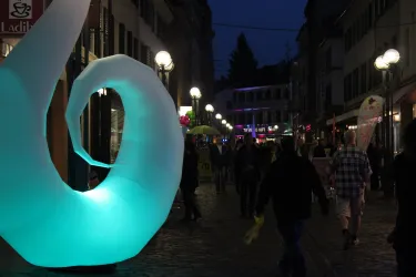Eine belebte Straße bei Nacht mit passierenden Menschen. Im Vordergrund steht eine große, leuchtende Skulptur in Form einer Spirale.