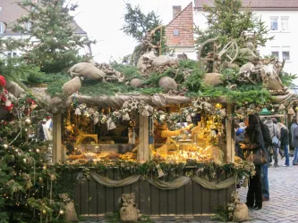 Ein festlich dekorierter Weihnachtsstand mit Lichtern und Tannenzweigen. Menschen schlendern um den Stand herum.