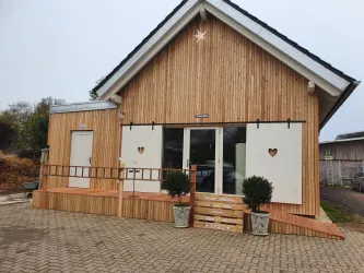 Ein stilvolles Holzhaus mit einem hellen, einladenden Eingangsbereich. Die Fassade ist mit Holzverkleidung versehen und hat dekorative Fensterläden mit Herzchen.