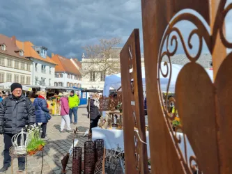 Ein belebter Markt mit vielen Ständen und Menschen. Im Vordergrund sind kunstvolle Metallherzen und im Hintergrund sind historische Gebäude zu sehen.