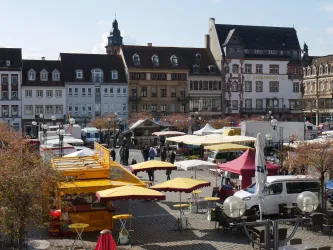 Ein belebter Marktplatz mit bunten Ständen und Sonnenschirmen. Im Hintergrund sind historische Gebäude und ein blauer Himmel zu sehen.