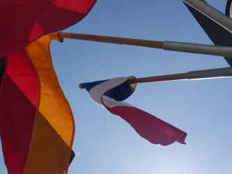 Drei Fahnen wehen im Wind vor einem klaren blauen Himmel. Die Fahnen zeigen die Farben Deutschland, Frankreich und möglicherweise eine weitere Nation.