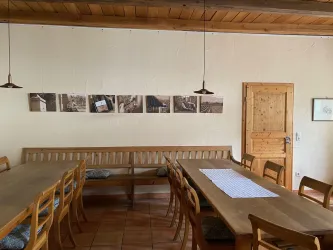 Ein heller Raum mit einem langen Holztisch und Stühlen. An der Wand hängen einige Bilder und eine Holztür führt nach draußen.