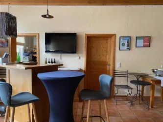 Ein gemütlicher Innenraum mit einer Bar und verschiedenen Sitzmöbeln. Die Wände sind hell gestrichen und es gibt eine TV-Leinwand.