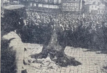 Eine Menschenmenge versammelt sich auf einem Platz. Im Vordergrund brennt ein Feuer auf dem Boden.