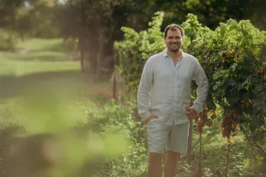 Ein Mann steht lächelnd in einem Weinberg. Im Hintergrund sind grüne Weinreben und eine sonnige Landschaft zu sehen.