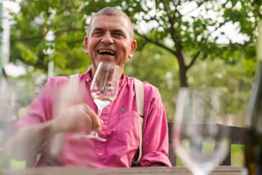 Ein fröhlicher Mann mit einem Glas in der Hand sitzt draußen und lacht. Im Hintergrund sind grüne Bäume zu sehen.
