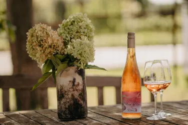 Eine Flasche Roséwein steht auf einem Holztisch, neben zwei Weingläsern. Im Hintergrund befindet sich eine Vase mit blühenden Hortensien.