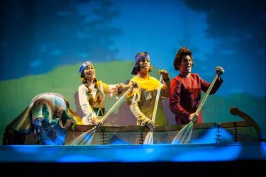 Eine Gruppe von Darstellern in farbenfrohen Kostümen sitzt in einem Boot und rudert. Im Hintergrund ist eine malerische Landschaft mit Bäumen und Blau zu sehen.