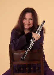 Eine lächelnde Frau mit langen Haaren sitzt auf einem Stuhl und hält ein Oboe. Sie trägt ein lila Oberteil und hat einen freundlichen Ausdruck.