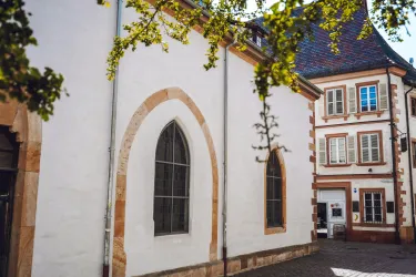 Eine historische Gebäudeansicht mit einem weißen Wand, einem großen, gotischen Fenster und einem Nachbargebäude aus Stein. Die Szene ist von sanften Baumzweigen umrahmt.
