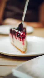Ein Stück Käsekuchen mit einer Fruchtschicht aus roter Marmelade auf einem weißen Teller. Ein Löffel ist in den Kuchen stecken.