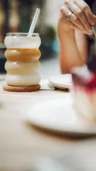 Ein Glas mit einem kühlen Getränk und einem Strohhalm steht auf dem Tisch. Im Hintergrund ist ein Stück Kuchen zu sehen.