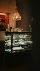Ein modernes Café mit einer Vitrine voller leckerer Torten und Gebäck. Sanftes Licht und Holzleisten schaffen eine einladende Atmosphäre.