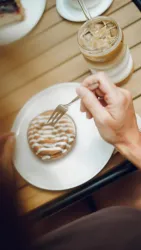 Eine Hand hält eine Gabel, während sie sich auf einen Keks mit Zuckerguss vorbereitet. Neben dem Keks steht ein Glas mit kaltem Kaffee.