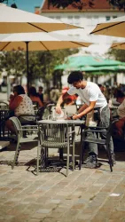 Ein junger Mensch deckt einen Tisch in einem Café im Freien. Im Hintergrund sieht man viele Gäste unter Sonnenschirmen.