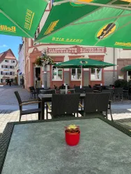 Ein gemütlicher Außenbereich eines Restaurants mit Tischen und Sonnenschirmen. Im Vordergrund steht ein Blumentopf auf einem Tisch.