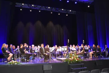 Ein Orchester sitzt auf der Bühne mit verschiedenen Instrumenten. Die Beleuchtung ist dunkelblau und es gibt Blumenarrangements im Vordergrund.