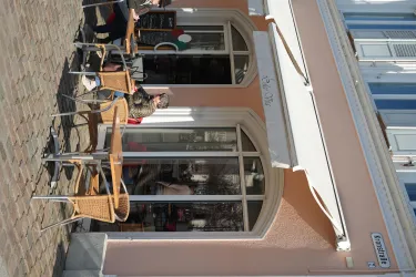 Eine gemütliche Caféterrasse mit Tischen und Stühlen. Im Hintergrund sieht man ein pinkes Gebäude und Gäste, die draußen sitzen.