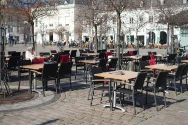 Ein leerer Platz mit zahlreichen Tischen und Stühlen im Freien. Im Hintergrund sind einige Gebäude und Bäume zu sehen.