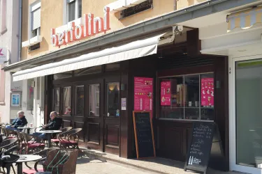 Ein gemütliches Restaurant mit einer einladenden Terrassenfläche. Die Fassade ist in dunklem Holz gehalten und es gibt ein großes Schaufenster mit einer Speisekarte.