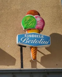 Ein bunter Eisschirm mit verschiedenen Eissorten in einer Waffel. Das Schild gehört zur Eisdiele Bertolini.