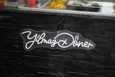 Ein Schild mit der Aufschrift „Yilmaz Döner“ ist an einer Theke angebracht. Der Hintergrund besteht aus dunklen Steinplatten.