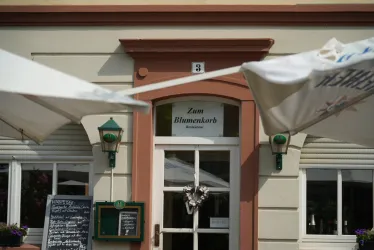 Ein gemütliches Restaurant mit dem Namen "Zum Blumenkorb". Der Eingangsbereich ist freundlich gestaltet und weist auf Speiseangebote hin.