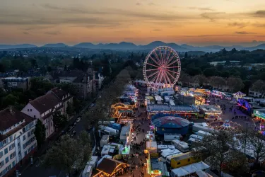 Ein buntes Volksfest bei Sonnenuntergang mit einem großen Riesenrad. Stände und Lichter prägen die Szene und sorgen für festliche Stimmung.