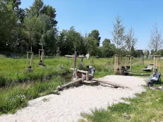 Ein Spielplatz am Wasser mit Holzgeräten und einer sandigen Uferzone. Im Hintergrund sind Bäume und Wiese zu sehen.