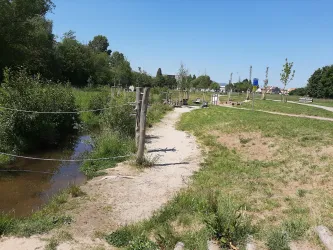 Ein ruhiger Weg neben einem kleinen Bach und grünem Gras. Im Hintergrund sind vereinzelte Bänke und Bäume sichtbar.