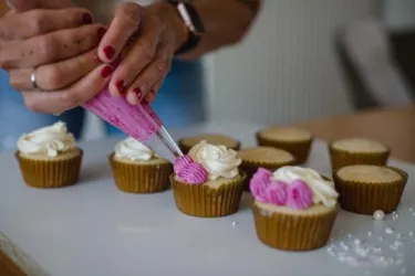 Zwei Hände spritzen rosa und weiße Buttercreme auf frisch gebackene Cupcakes. Im Hintergrund sind weitere Cupcakes zu sehen, die bereits verziert sind.