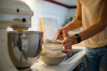 Eine Person sieben Mehl in eine Schüssel in einer modernen Küche. Im Hintergrund steht eine Küchenmaschine auf der Arbeitsplatte.