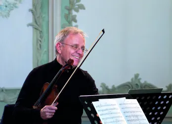 Ein lächelnder Geiger hält seine Violine und steht vor Notenblättern. Der Hintergrund zeigt eine elegante Wandgestaltung.
