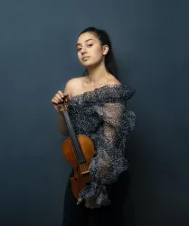 Eine junge Frau steht vor einem blauen Hintergrund und hält eine Violine in der Hand. Sie trägt ein auffälliges, gemustertes Oberteil und hat ihre Haare zurückgebunden.