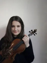 Eine junge Frau steht vor einem blauen Hintergrund und hält eine Violine in der Hand. Sie trägt ein auffälliges, gemustertes Oberteil und hat ihre Haare zurückgebunden.