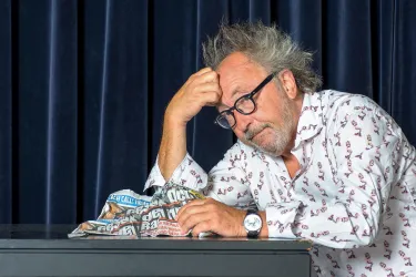 Ein nachdenklicher Mann mit zerzaustem Haar sitzt an einem Tisch und hält eine zusammengeknüllte Zeitung in der Hand. Er sieht besorgt oder frustriert aus.