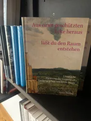 Ein Buch mit dem Titel "Aus einer geschützten Ecke heraus lässt du den Raum entstehen" steht auf einem Regal. Es sind weitere Bücher in verschiedenen Farben im Hintergrund sichtbar.