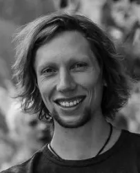 Ein junger Mann mit lockigem Haar und lächelndem Gesicht. Der Hintergrund ist unscharf und zeigt eine natürliche Umgebung.