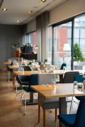 Ein modernes Restaurant mit Holztischen und farbigen Stühlen. Große Fenster sorgen für viel Tageslicht und Blick auf die Terrasse.