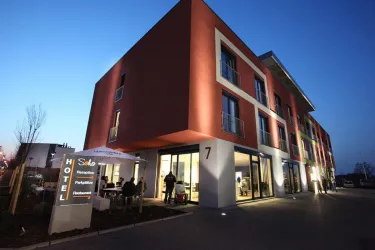 Ein modernes Hotelgebäude bei Dämmerungslicht. Vor dem Hotel befinden sich Gäste, die auf der Terrasse sitzen.