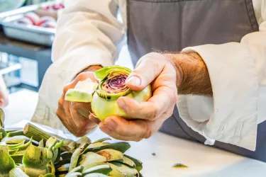 Eine Person schält eine Artischocke. Im Hintergrund sind weitere Artischocken zu sehen.